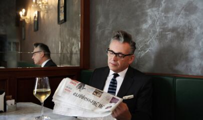 Götz Alsmann an einem Tisch im Restaurant mit Zeitung in der Hand und Glas Wein vor sich. Link führt zum Kalendereintrag